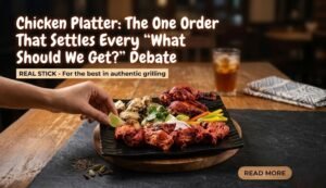 Chicken platter on a wooden board with grilled chicken, vegetables, and a hand reaching to pick a piece; coffee tab and napkin in the background.