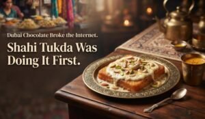 Plate of Shahi Tukda topped with cream and nuts on an ornate plate, with a brass teapot and cup on a wooden table; blurred market setting in the background.